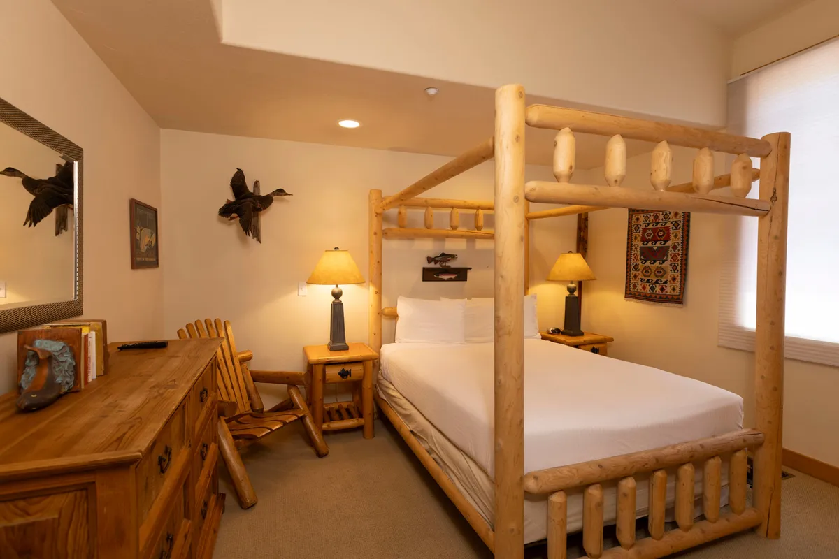 This image showcases a serene bedroom with a four-poster bed dressed in soft linens, complemented by a wooden dresser, table lamps, and decorative artwork. The space is bathed in natural light, creating a peaceful atmosphere ideal for relaxation and rejuvenation.