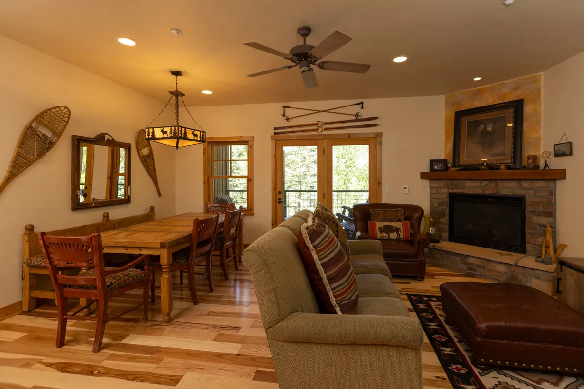 An inviting dining area seamlessly connected to the living space, featuring a wooden dining table positioned near a glowing fireplace. The seating area is visible beyond, suggesting an open-plan layout. The large windows flood the space with natural light and offer glimpses of the outdoors.