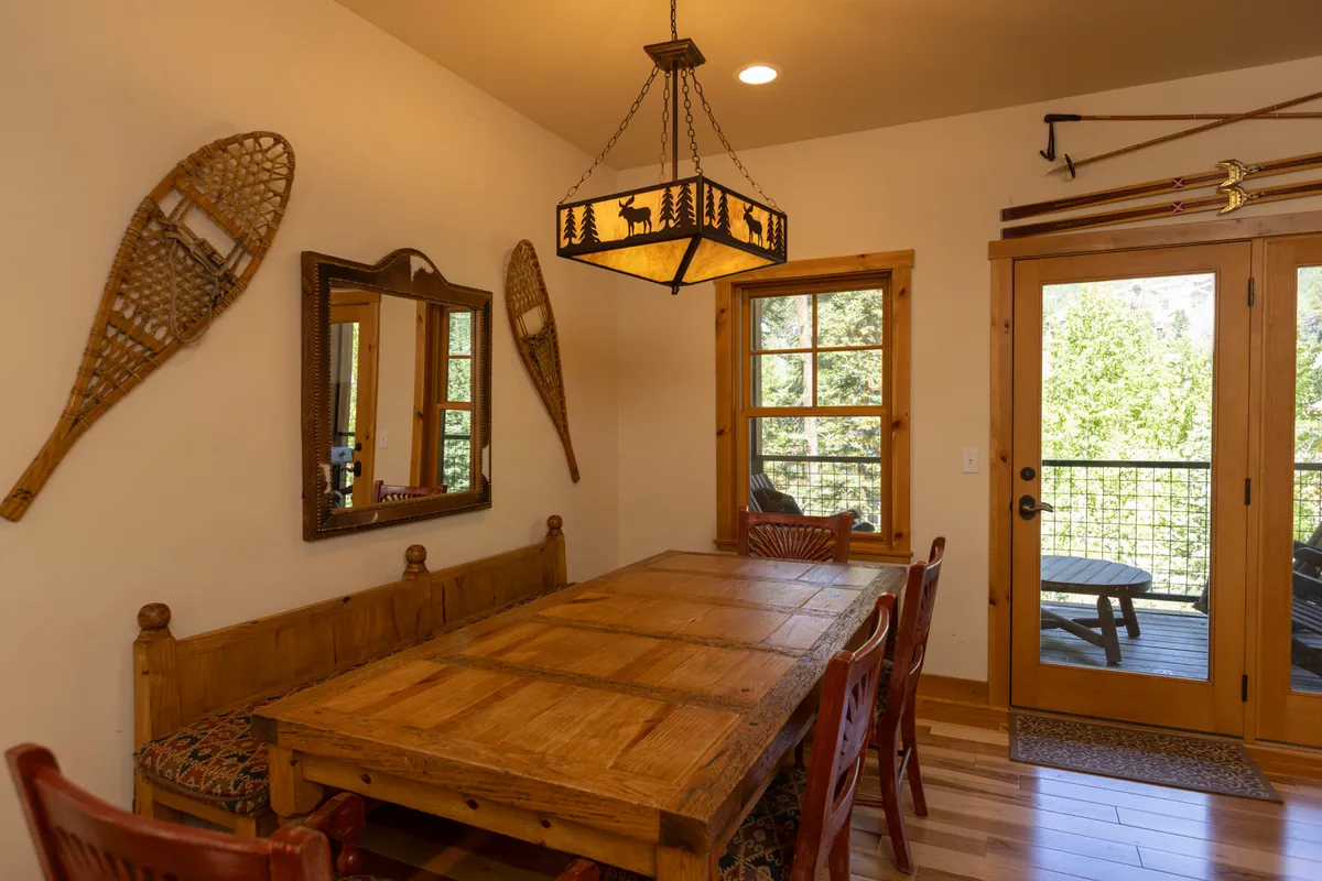 A warm, welcoming dining area with a sturdy wooden dining table and a nature-inspired chandelier overhead. Large windows on one side allow plenty of sunlight in and hint at beautiful outdoor views. The setup is perfect for family meals, gatherings, or board games on cozy evenings.