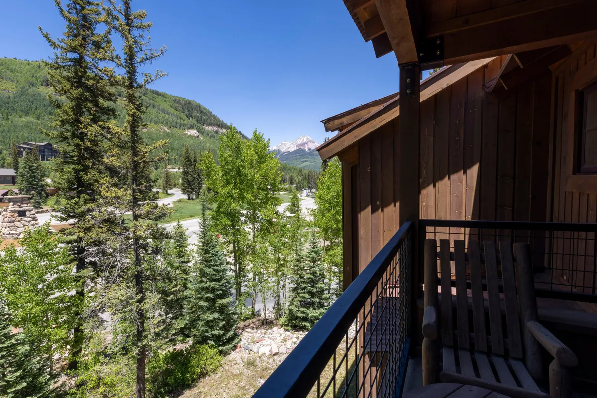A wooden balcony set against a backdrop of lush green trees and distant mountain peaks. The space invites relaxation, offering a peaceful spot for stargazing, sipping coffee, or soaking in the panoramic scenery with easy access to the outdoors.