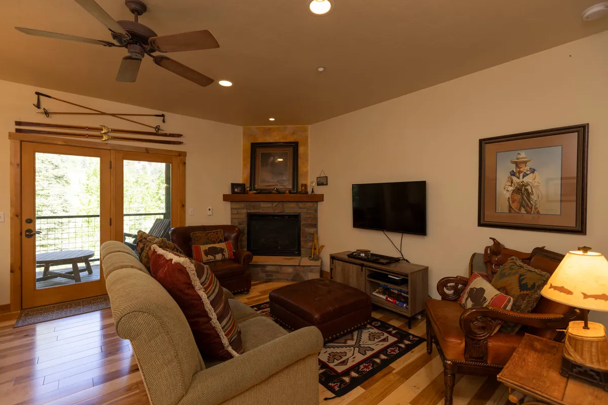 A spacious open-plan living area featuring a striking stone fireplace as the focal point. Large windows in the background frame scenic mountain views, while cozy, plush seating—including a sofa and armchairs—creates an inviting space for gatherings. Artwork on the walls and thoughtful lighting enhance the warm, welcoming atmosphere.