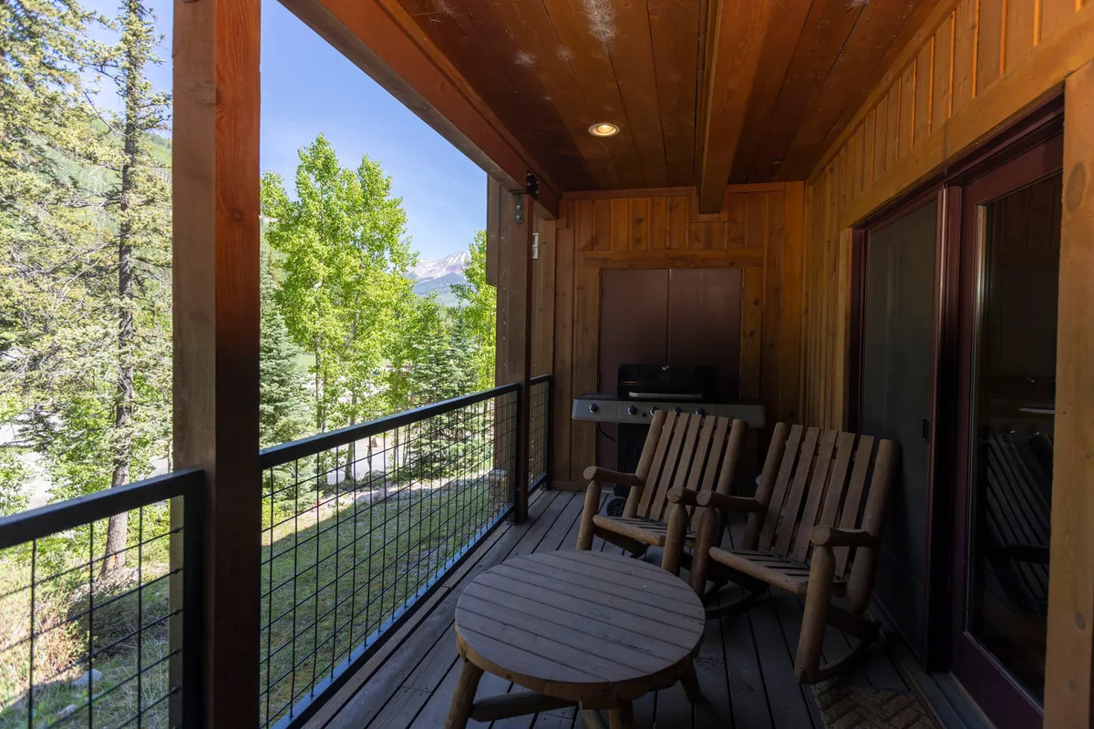 A spacious wooden deck furnished with rocking chairs overlooks a serene forest and offers a sweeping view of the surrounding mountains. The deck is bathed in natural light, perfect for enjoying a coffee at sunrise, reading, or relaxing after a day of adventure.