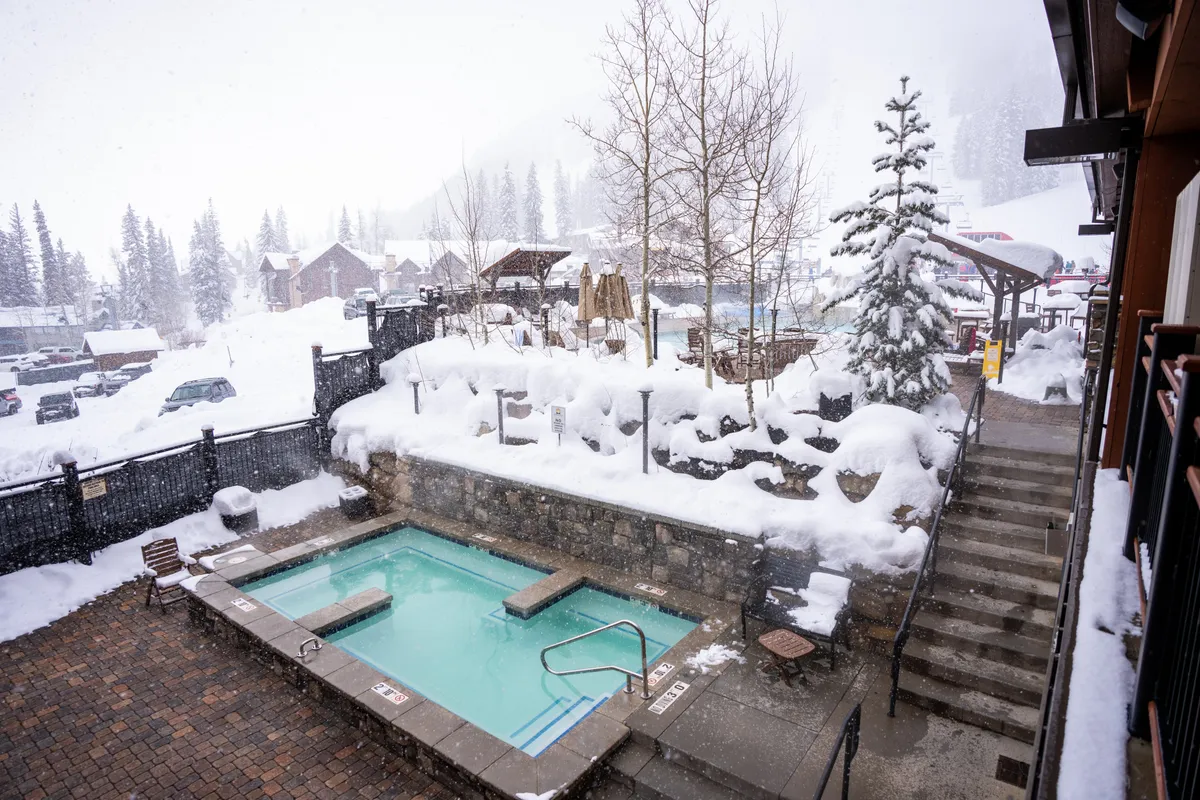 A serene outdoor hot tub set amidst a snowy landscape and encircled by tall trees. Stepping stones lead the way, highlighting the cozy contrast between the warm water and the crisp mountain air. The setting suggests a tranquil, social experience surrounded by nature’s beauty.