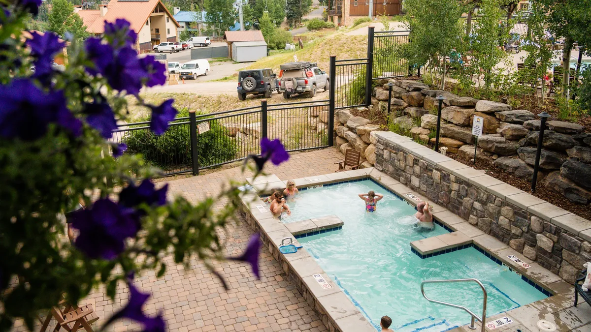 A lively exterior shot featuring a sparkling swimming pool surrounded by lush landscaping and blooming flowers. Several people are seen enjoying the pool and sunbathing, with the mountains faintly visible in the background, suggesting an inviting communal space for relaxation and fun across the seasons.