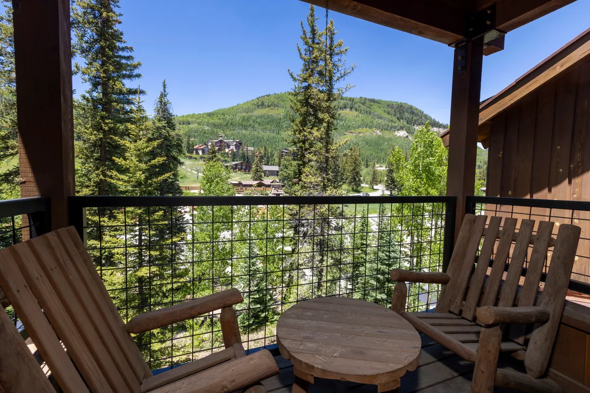 The image showcases a private balcony at the condo, furnished with inviting wooden chairs and a small round table, all set against a backdrop of majestic mountain views and towering pine trees. The peaceful scene suggests opportunities for relaxing, sipping coffee, or simply soaking in the natural beauty.