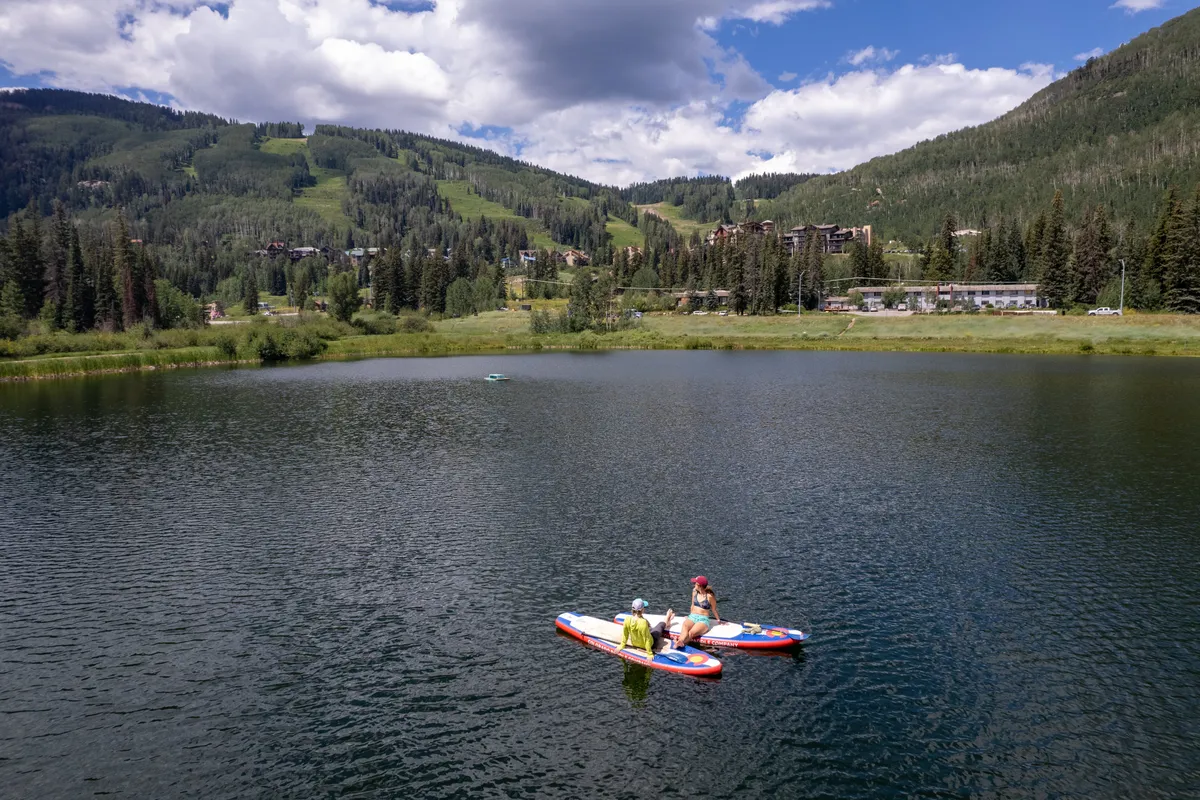 A tranquil mountain lake scene with paddleboarders gliding across calm waters, framed by dramatic mountains and a cloudy sky. The natural setting is peaceful and open, suggesting endless opportunities for outdoor adventure and relaxation.