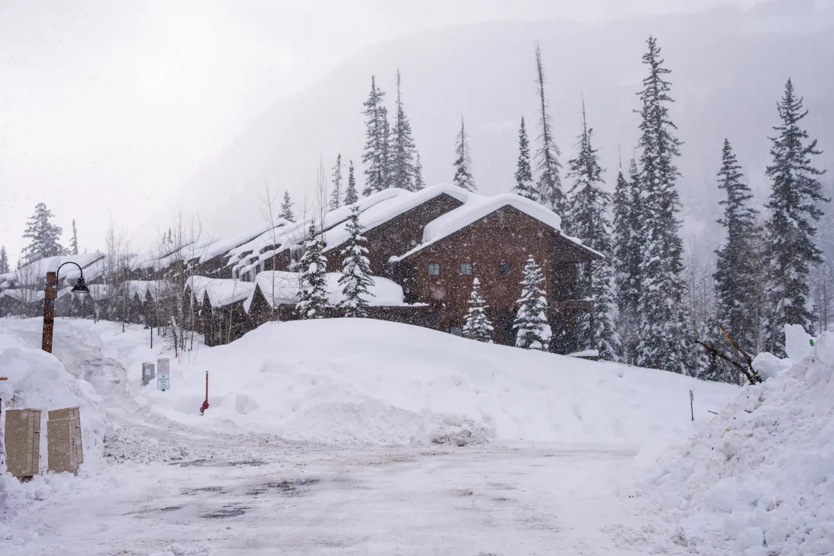 Charming, snow-dusted buildings surrounded by tall pine trees create a cozy, inviting winter village ambiance. A lamp post softly glows against the backdrop of falling snow, evoking après-ski relaxation and community.
