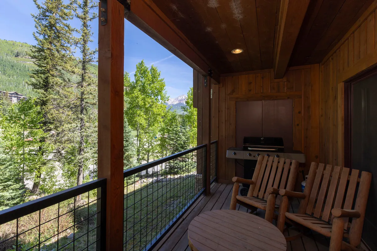 The exterior setting features wooden rocking chairs on a patio, an outdoor grill, and a sweeping view of surrounding trees and mountains. The scene invites relaxation after a day outdoors, with opportunities for outdoor dining and soaking in the mountain scenery.