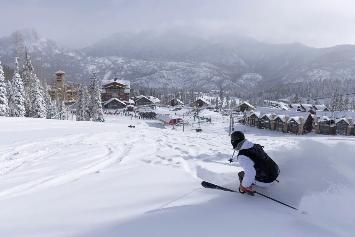 A skier in motion glides down freshly groomed slopes with a snow-covered mountain backdrop. The image includes visible ski resort buildings and a ski lift, creating an immersive sense of being at the heart of a vibrant winter playground.
