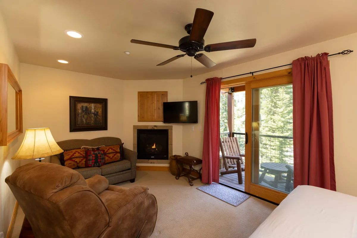 An inviting interior living area that opens directly onto a balcony. The space features a cozy sofa, a warm fireplace, and an armchair positioned for views through large glass doors to the balcony and mountains beyond, highlighting the easy transition between indoors and outdoors.
